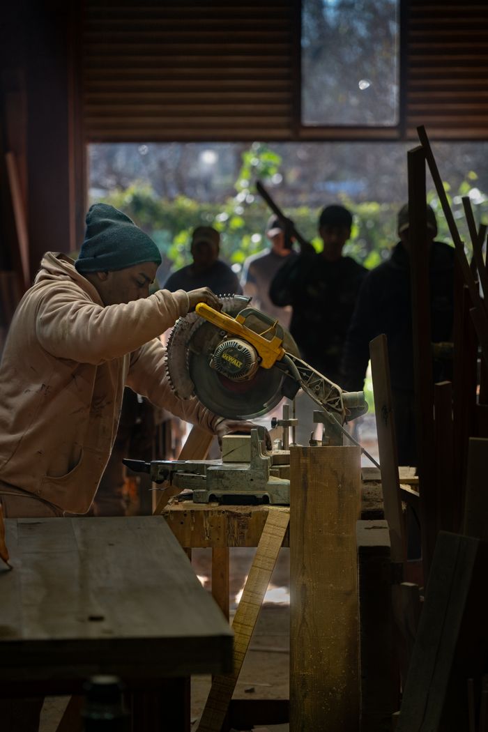 A woodworker focuses intently on using a circular saw in a sunlit workshop, surrounded by colleagues.